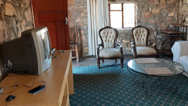 Een kamer met een stenen muur, een houten bureau met een computer en telefoon, twee fauteuils, een glazen salontafel en een witte bank met rode kussens.
