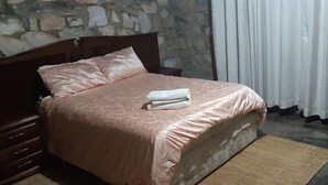Een bed met een roze sprei en een witte handdoek, een houten nachtkastje, een stenen muur en een gordijn.
