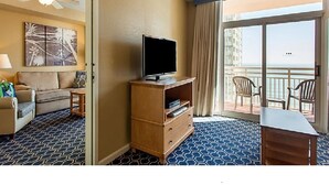 Habitación de hotel con televisor sobre un soporte de madera, sofá, mesa de comedor y balcón con sillas.