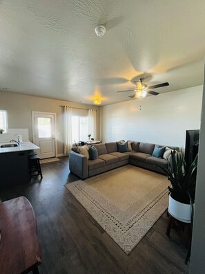 A living room with a sectional sofa, a ceiling fan, a kitchen area with a sink, and a dining area with a table and chairs.