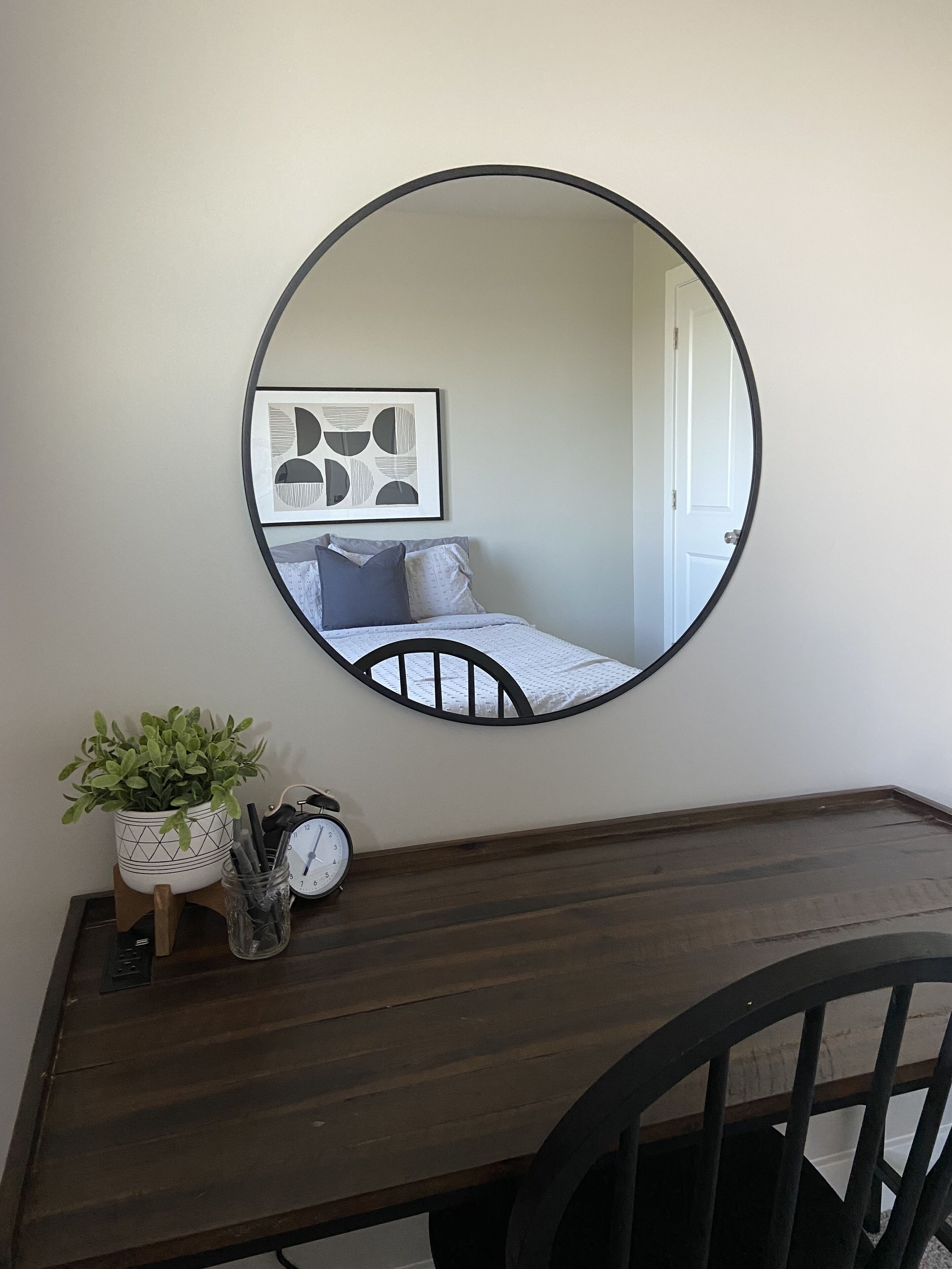 A round mirror on a wall, a bed with a headboard, a bedside table with a clock, a potted plant, and a framed picture.