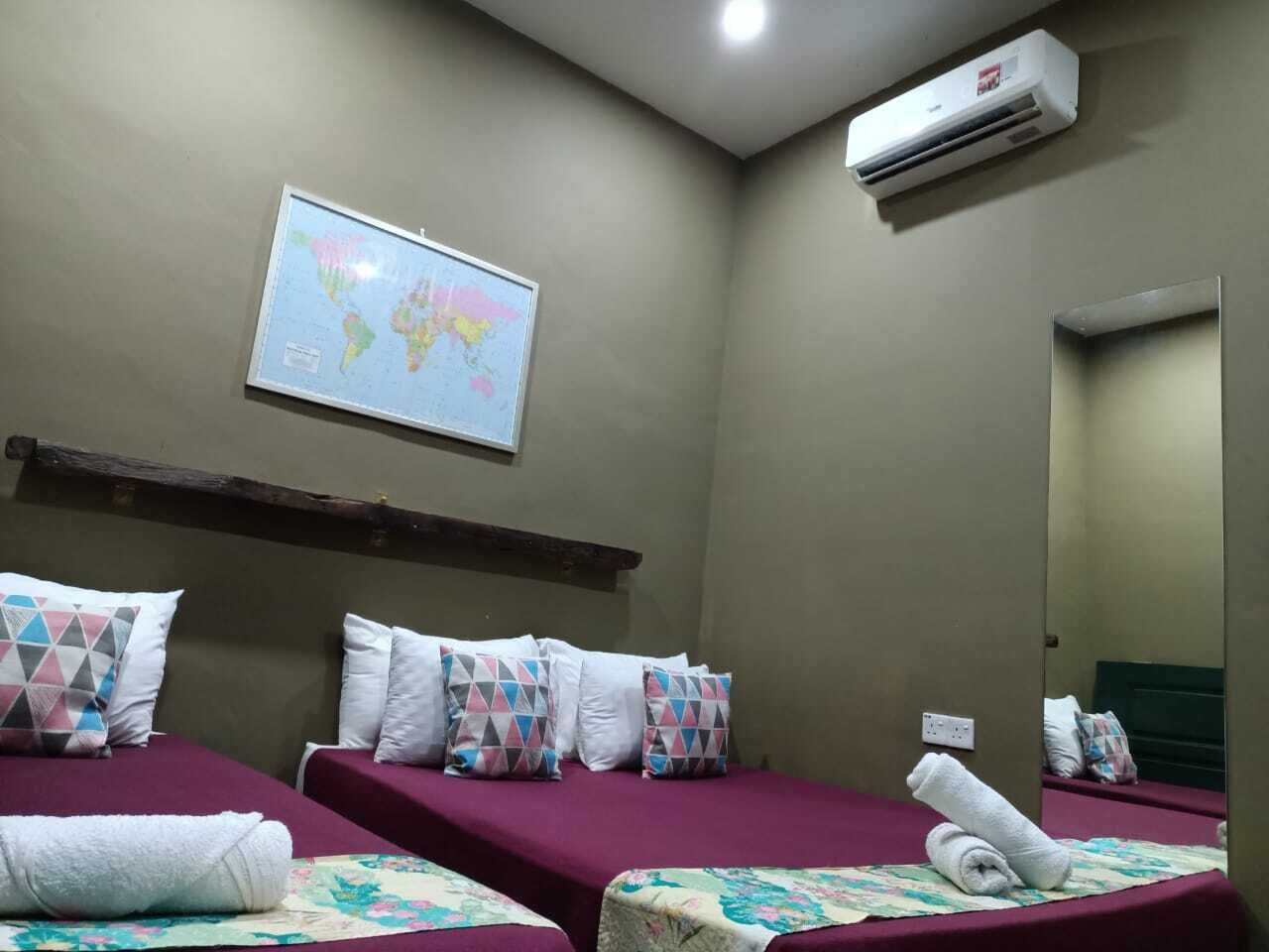 Habitación de hotel con dos camas, cada una con cubrecama morado y almohadas blancas. Hay un mapa mundial en la pared y un equipo de aire acondicionado sobre las camas.