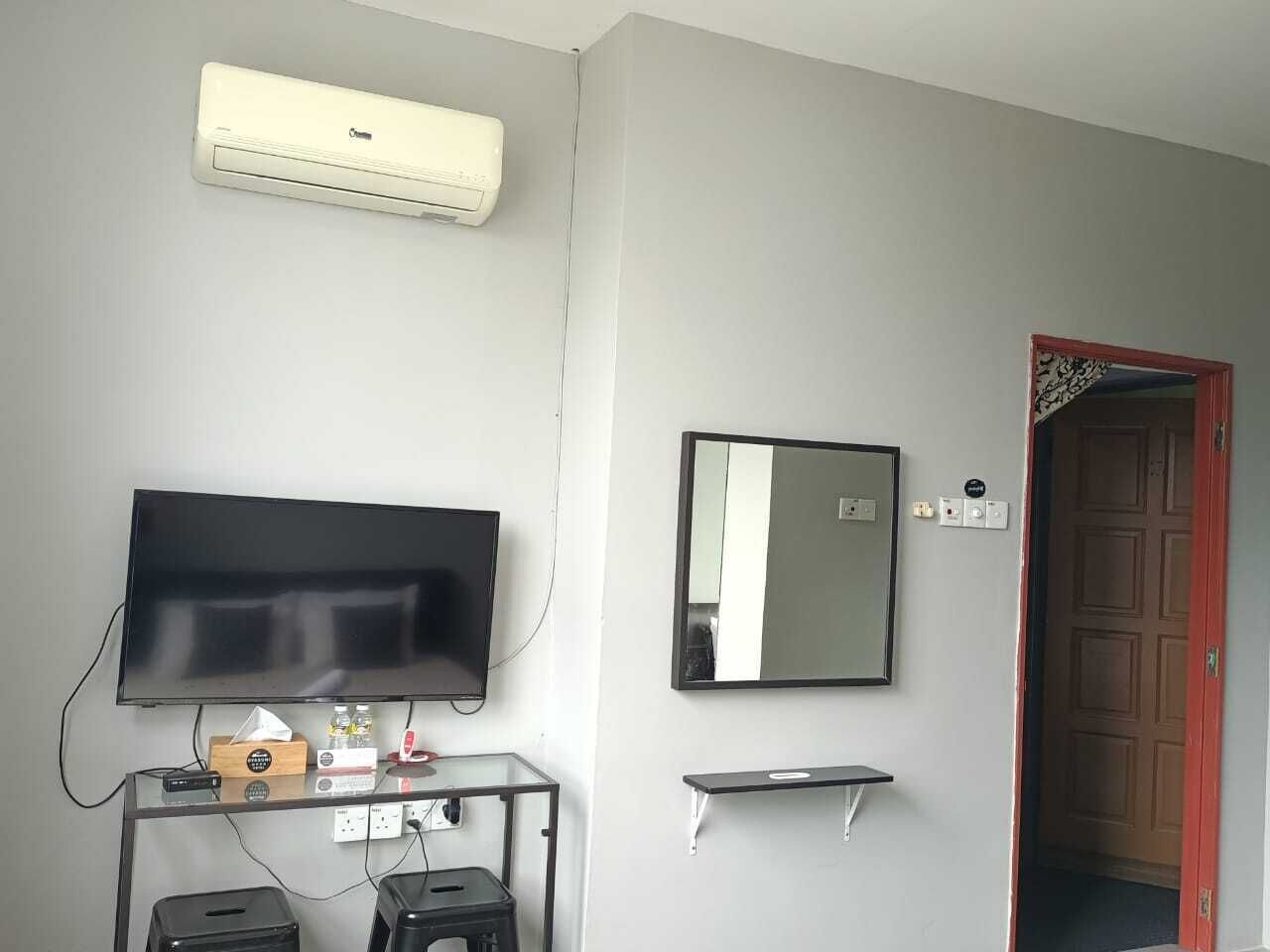 Una habitación con una televisión de pantalla plana, un espejo adosado a la pared, un mueble de televisión de madera con un control remoto, un estante pequeño y una puerta con marco rojo.