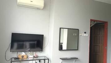Una habitación con una televisión de pantalla plana, un espejo adosado a la pared, un mueble de televisión de madera con un control remoto, un estante pequeño y una puerta con marco rojo.