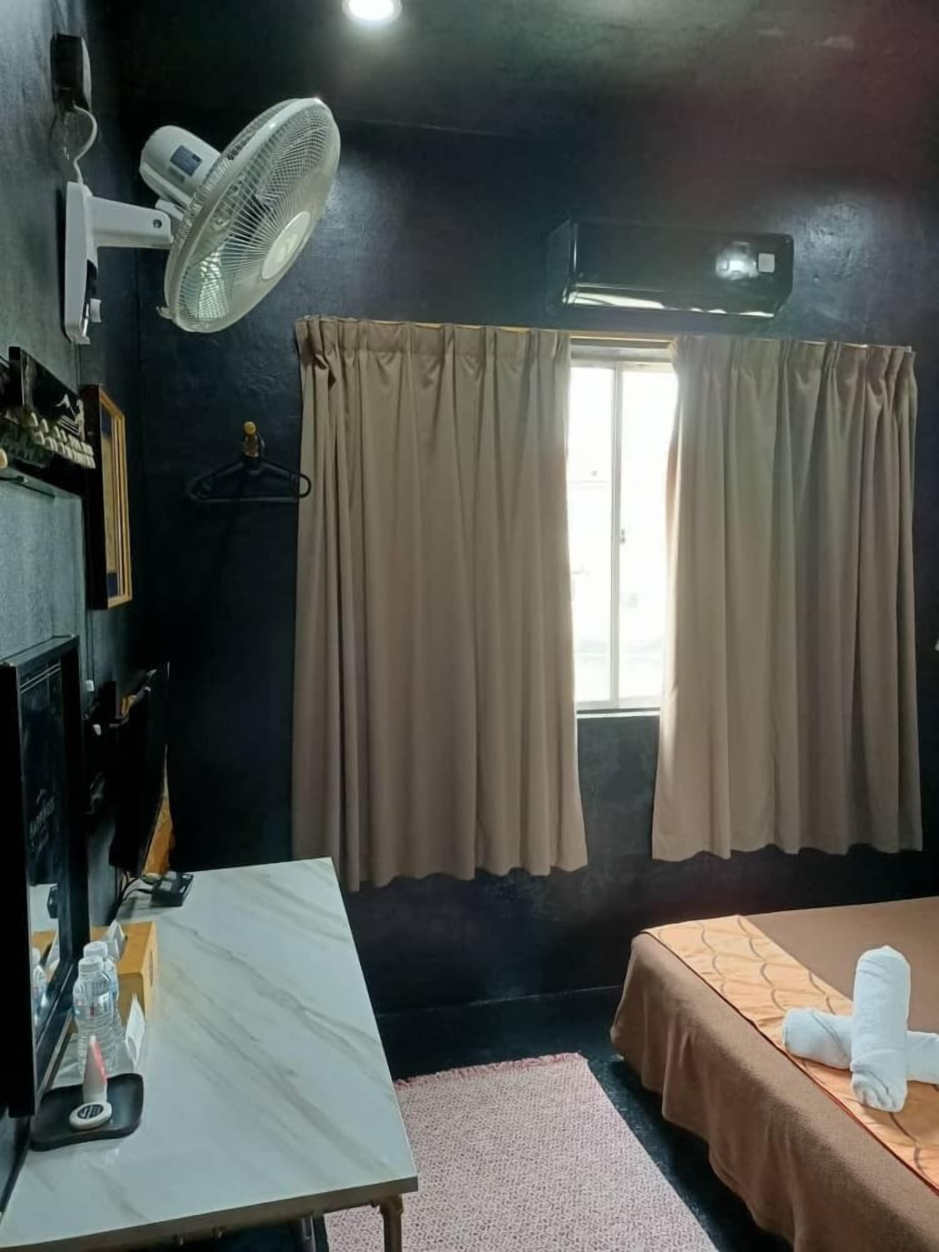 Une chambre d’hôtel équipée d’un lit, d’une table, d’un ventilateur, d’une fenêtre avec des rideaux et d’un climatiseur fixé au mur.