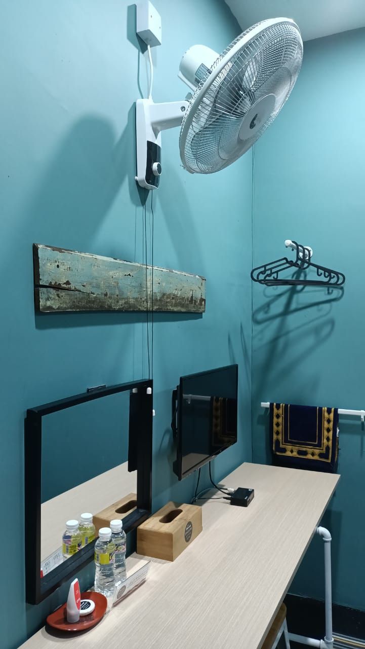 Una habitación con un escritorio, dos monitores, un ventilador, una estantería de madera y un perchero.