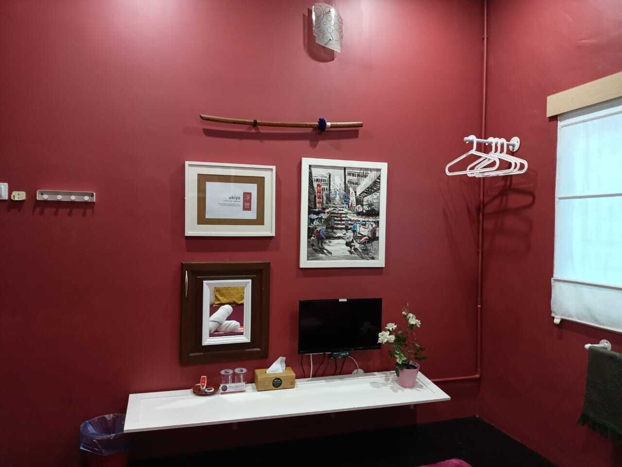 A room with a red wall, a white desk with a flat-screen TV, framed pictures, a small plant, and a towel rack.