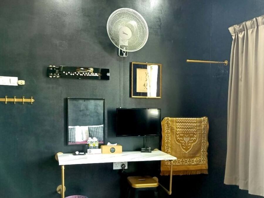 A room with a dark wall, a white desk with a computer, a fan, a framed picture, a towel, and a curtain.