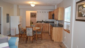 A compact kitchen with wooden cabinets, a refrigerator, a dining table with chairs, and a window with blinds. - Cattaraugus, Your home away from home (Cattaraugus)