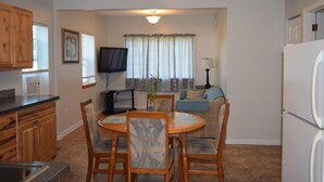 A compact kitchen with wooden cabinets, a dining table with chairs, a television, and a window with curtains. - Cattaraugus, Your home away from home (Cattaraugus)