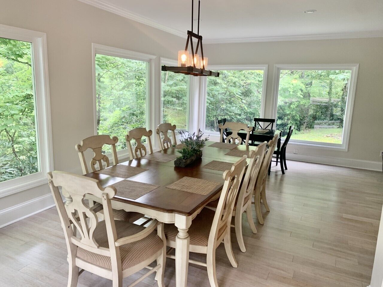 Une salle à manger avec une grande table en bois entourée de chaises, un lustre et de grandes fenêtres donnant sur un paysage verdoyant.
