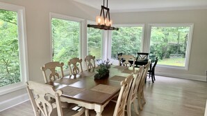 A dining room with a large wooden table surrounded by chairs, a chandelier, and large windows offering a view of greenery outside.