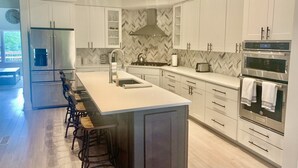 A modern kitchen with white cabinetry, a large island with bar stools, stainless steel appliances, and a herringbone-patterned backsplash.