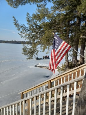 Terraza de madera con vista a un lago congelado, una bandera estadounidense, árboles y un vehículo para nieve a lo lejos.