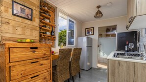 A kitchen with wooden cabinetry, a dining area with wicker chairs, a refrigerator, and a window with a view of greenery.