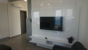 A modern living room with a flat-screen TV mounted on a white wall, a grey sofa, and a small plant on a shelf.