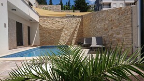 A swimming pool with a stone wall backdrop, a covered seating area, and a view of surrounding buildings.