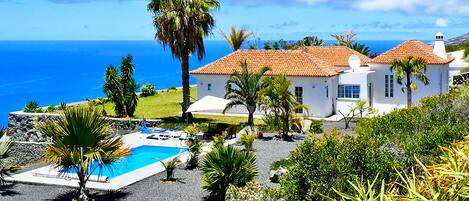 A coastal house with a swimming pool, surrounded by palm trees and a view of the sea.