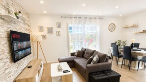 En moderne stue med en modulsofa, fjernsyn monteret på væggen, et spiseområde med bord og stole, og et stort vindue med gardiner.