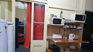 A compact kitchen with a microwave, toaster, and kettle on shelves, a refrigerator, and a red curtain. - Golden Mount Rest, 9 guest's max, 1 Double, 7 Singles  (Mount Morgan)