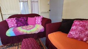 A room with a large, dark-colored sofa with a colorful tie-dye blanket and multiple pillows. There is a smaller, orange sofa with a pink floral cushion. The room has a simple, functional design with a plain door and a window with curtains. - Golden Mount Rest, 9 guest's max, 1 Double, 7 Singles  (Mount Morgan)