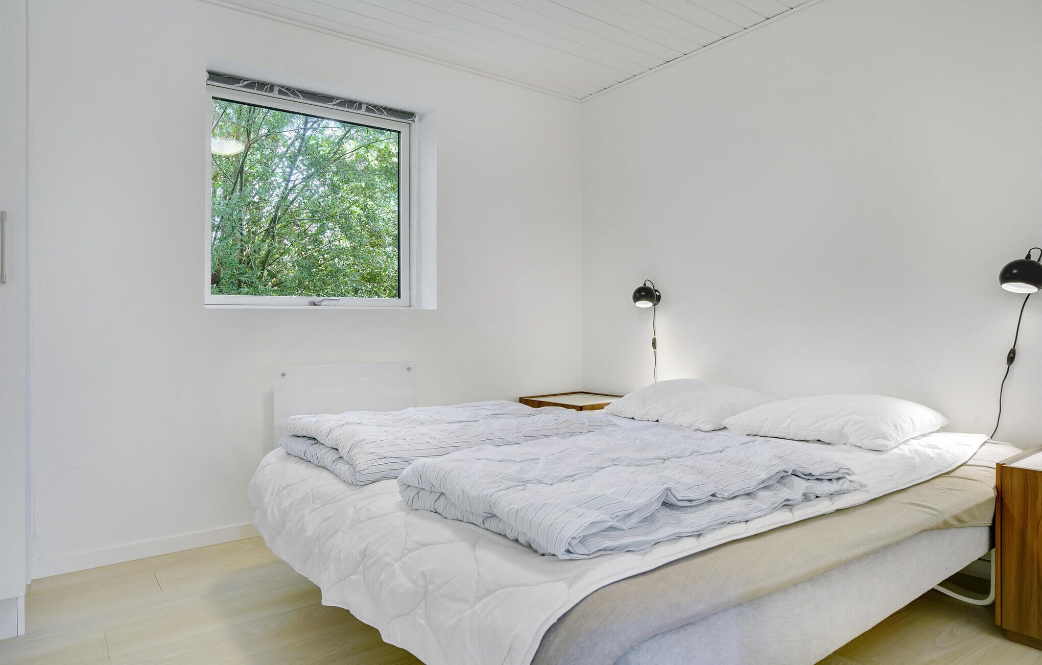 A minimalist bedroom with a large window, a bed with a white duvet and pillows, two bedside lamps, and a wooden nightstand.