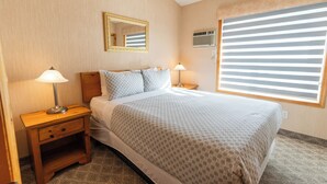 Una habitación de hotel ordenada con una cama, dos mesitas de noche con lámparas, una mesita auxiliar, un espejo y una ventana con persianas.