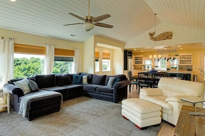 A living room with a sectional sofa, a white armchair, a ceiling fan, and a kitchen area visible through an open doorway.