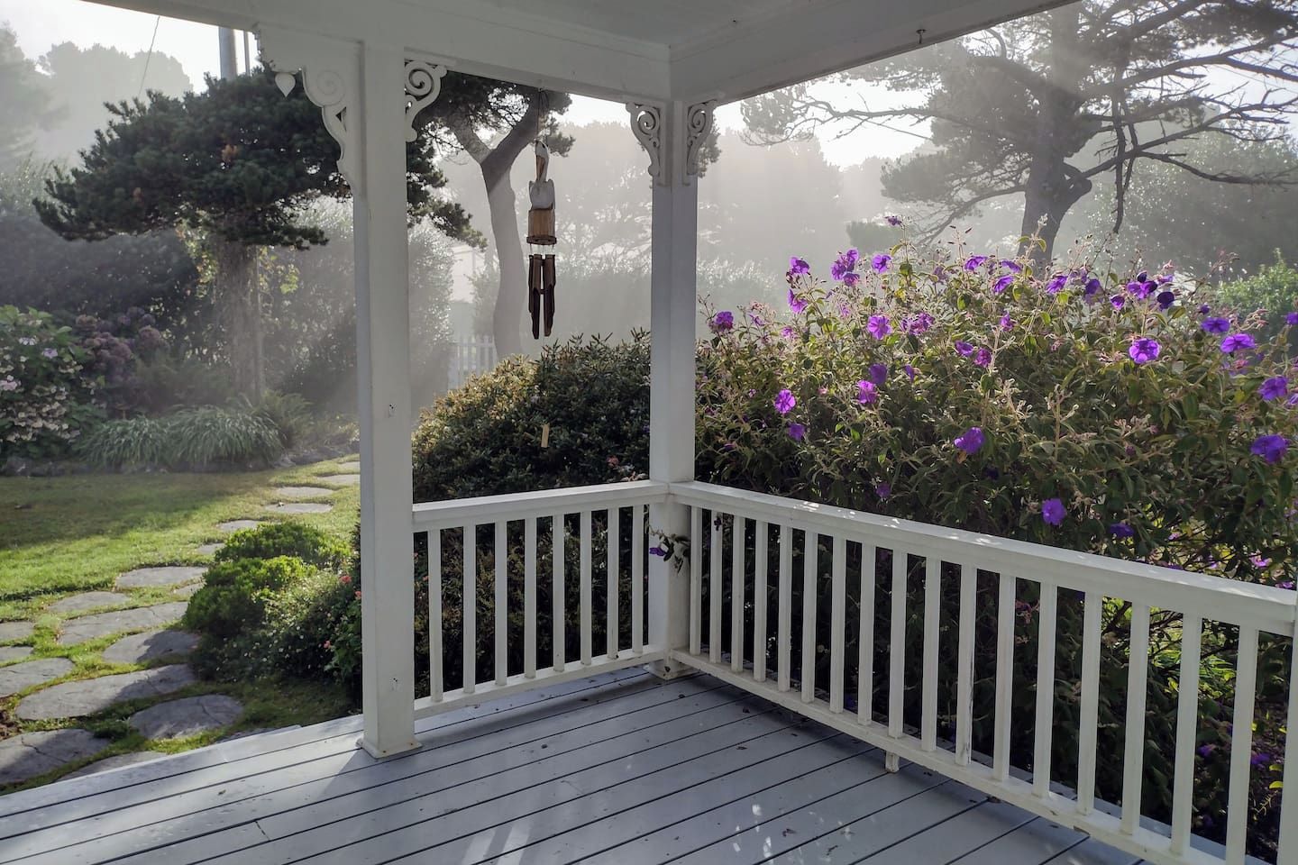 Een overdekt terras met een witte balustrade, een pad van stenen en een tuin met paarse bloemen.