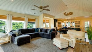 A spacious living room with a sectional sofa, a white armchair, and a wooden coffee table. There is a ceiling fan, a kitchen area with a dining table, and a view of greenery outside.