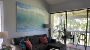 Ein Wohnzimmer mit großem Gemälde einer Brandungswelle, einer Couch mit Kissen, einem Couchtisch, einer Lampe und einer Glastür, die zu einem Außenbereich mit Tisch und Stühlen führt.