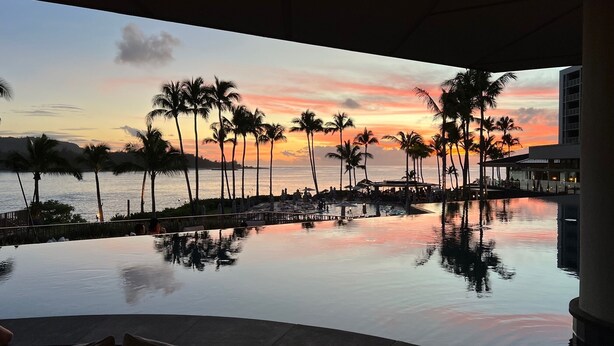 Blick auf ein Schwimmbecken bei Sonnenuntergang, mit Palmen und einer entfernten Küste.