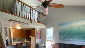 Ein geräumiger Wohnbereich mit Deckenventilator, großem Teppich, einer Couch mit Kissen und einem Wandbild von einer Welle.