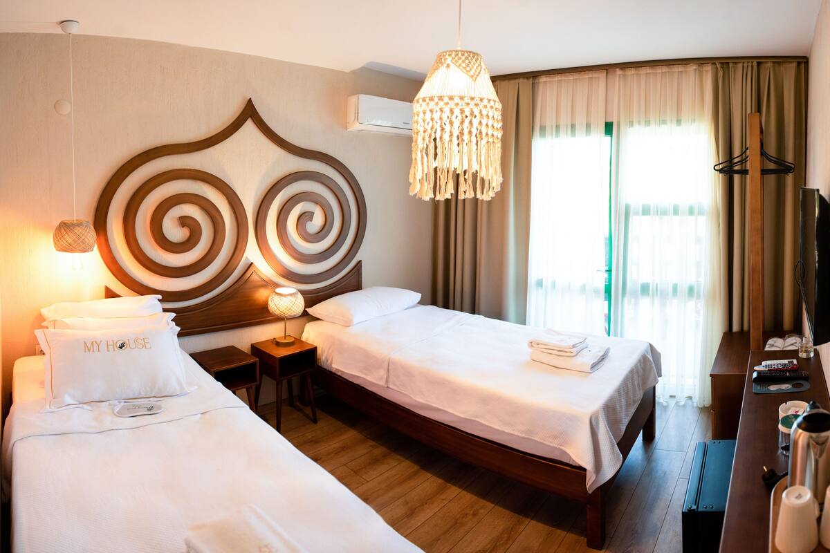 Hotelkamer met twee bedden, een houten hoofdbord, een hanglamp, een nachtkastje met lamp, een televisie en een raam met gordijnen.
