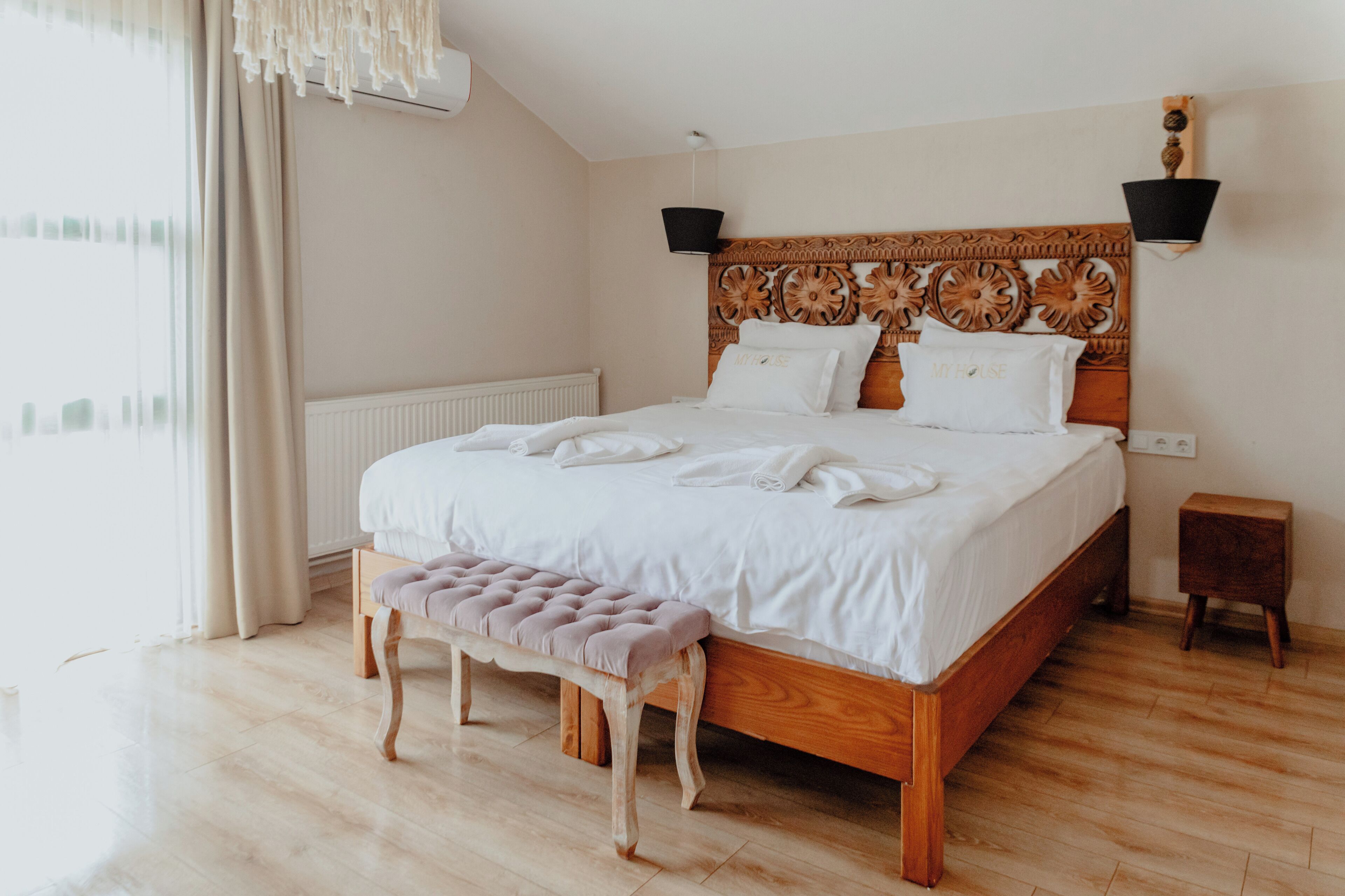 Una cama bien hecha con sábanas y almohadas blancas, un cabecero de madera con intrincados grabados, un banco al pie de la cama y una mesita de noche de madera al lado.