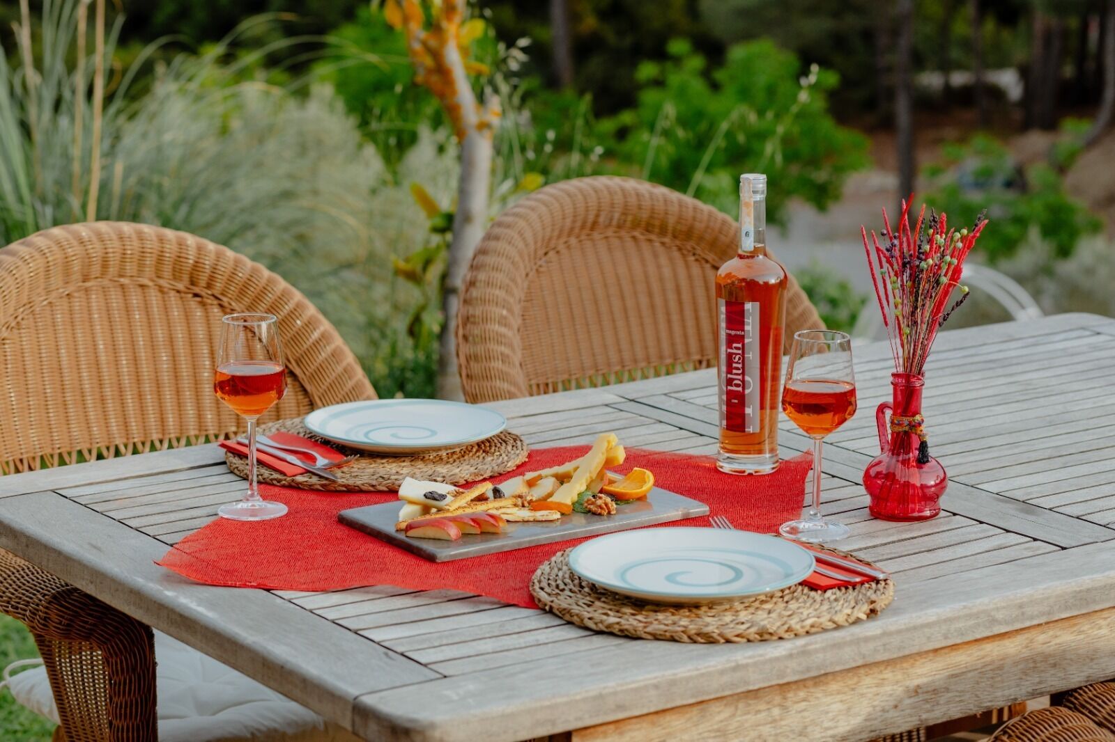 Una mesa de comedor al aire libre con sillas de mimbre, una botella de licor, vasos y una bandeja de frutas.