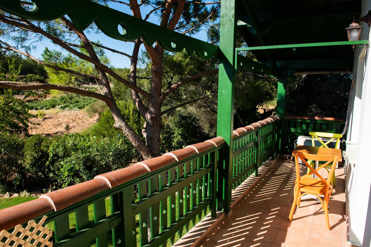 Een balkon met een groene balustrade, een houten stoel en uitzicht op bomen en heuvels.