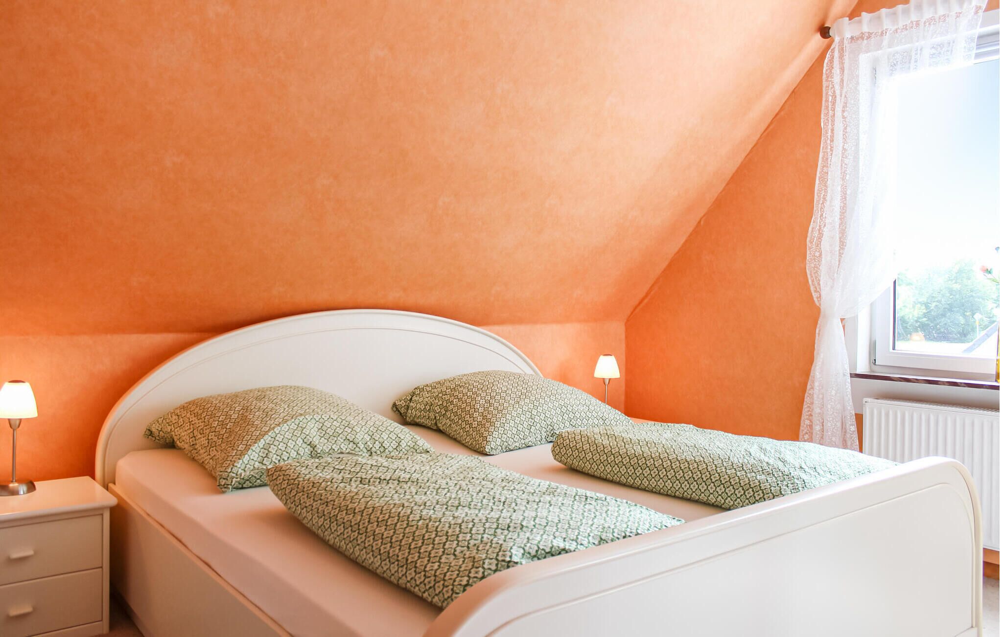 Ein Schlafzimmer mit einem weißen Bett, zwei Kissen, einem Nachttisch mit Lampe und einem Fenster mit Spitzenvorhängen.