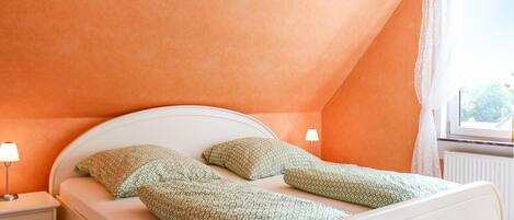 Ein Schlafzimmer mit einem weißen Bett, zwei Kissen, einem Nachttisch mit Lampe und einem Fenster mit Spitzenvorhängen.