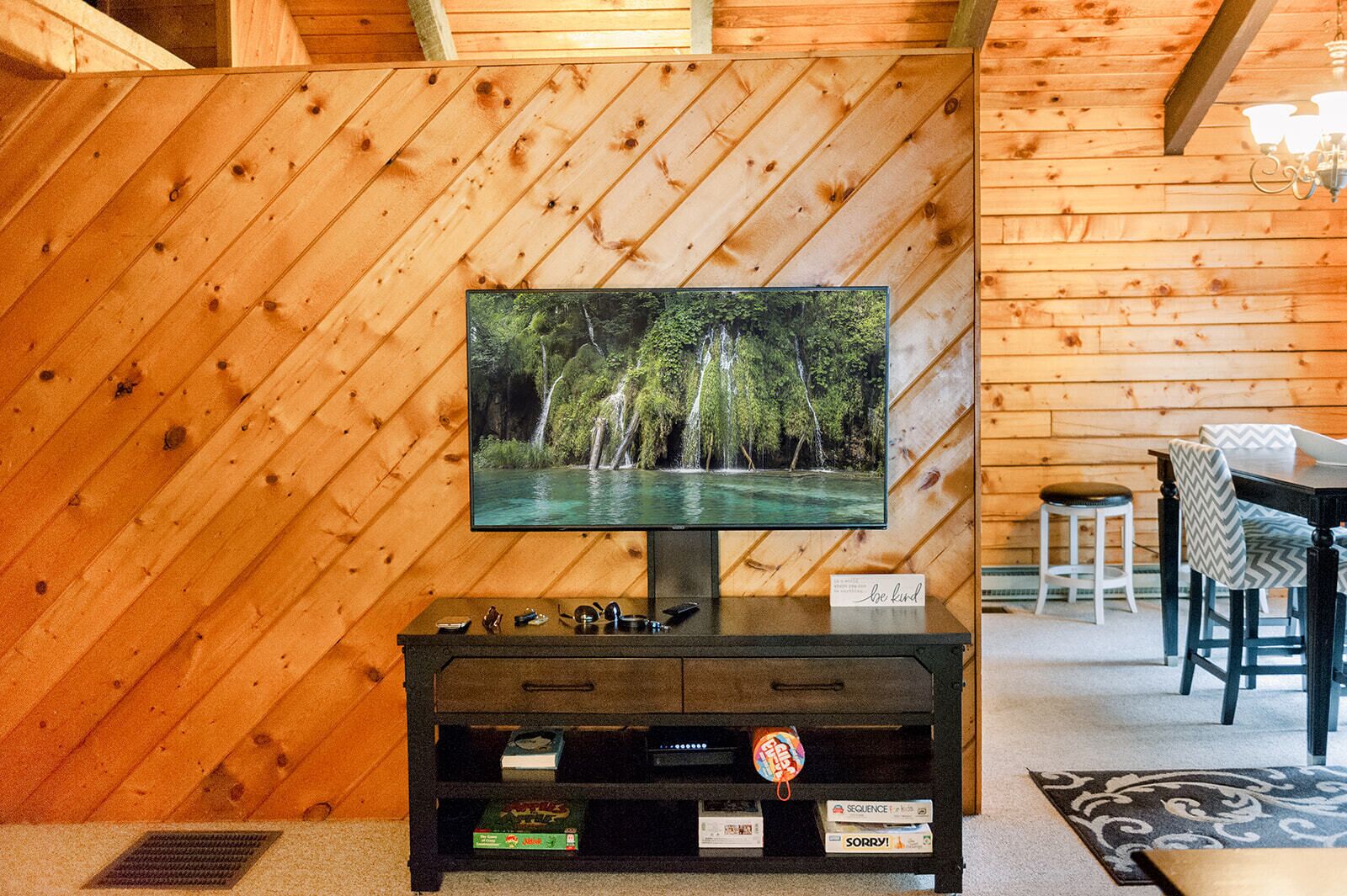 A television mounted on a wooden wall with a forest scene displayed, a small table with chairs, and a cabinet below the TV.