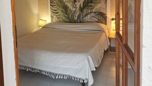 Une chambre à coucher comprenant un lit, une table de chevet, une lampe fixée au mur et une grande fresque murale ornée de motifs de feuilles tropicales.