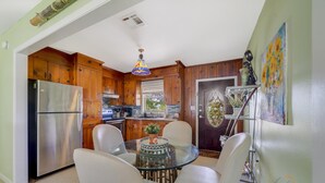 A kitchen with wooden cabinets, a stainless steel refrigerator, a glass dining table with four chairs, and a decorative ceiling light.