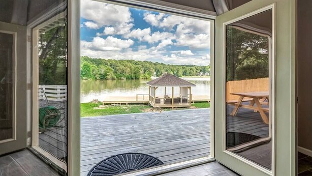 A view from inside a room with a wooden deck leading to a gazebo by a lake.