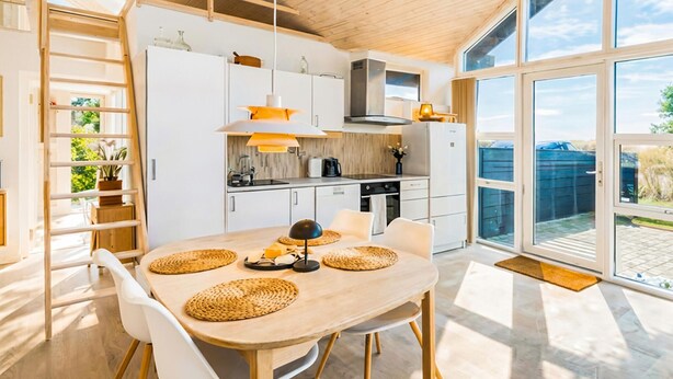 Eine moderne Küche mit weißen Schränken, einem Holztisch mit gewebten Tischsets und Blick ins Freie durch große Fenster.
