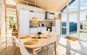 Une cuisine moderne avec des meubles blancs, une table à manger en bois agrémentée de sets de table en fibres naturelles, et une vue sur l’extérieur grâce à de grandes fenêtres.