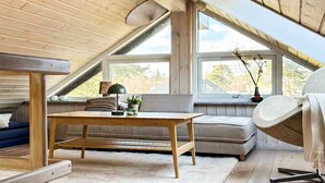 Ein gemütliches Zimmer im Dachgeschoss mit Holzbalkendecke, einer Couch, einem Tisch und großen Fenstern mit Blick ins Freie.