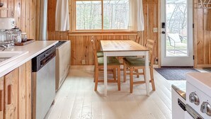 A wooden cabin kitchen with a dining table, chairs, a stove, and a window with curtains. - Cozy Vintage Cabin on Lake Pokegama & MTB Trails (Cohasset)