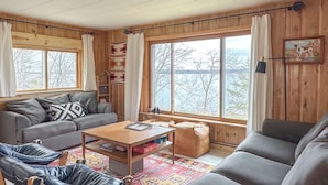 A cozy living room with a sofa, a coffee table, and a chair. There is a window with a view of trees and a lamp on a table. - Cozy Vintage Cabin on Lake Pokegama & MTB Trails (Cohasset)