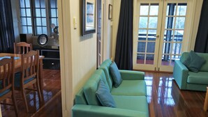 A living room with a green sofa, a wooden dining table with chairs, and a large window with curtains. - At the Centre of Gallery Walk, Tamborine Mountain (Eagle Heights)