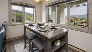 Een eethoek met een tafel voor vier personen, twee ramen met uitzicht op een landschap, en een kamerplant op de tafel.